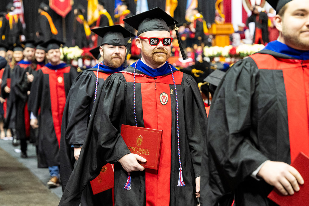 A man in a graduation gown wearing sunglasses walks across the stage.