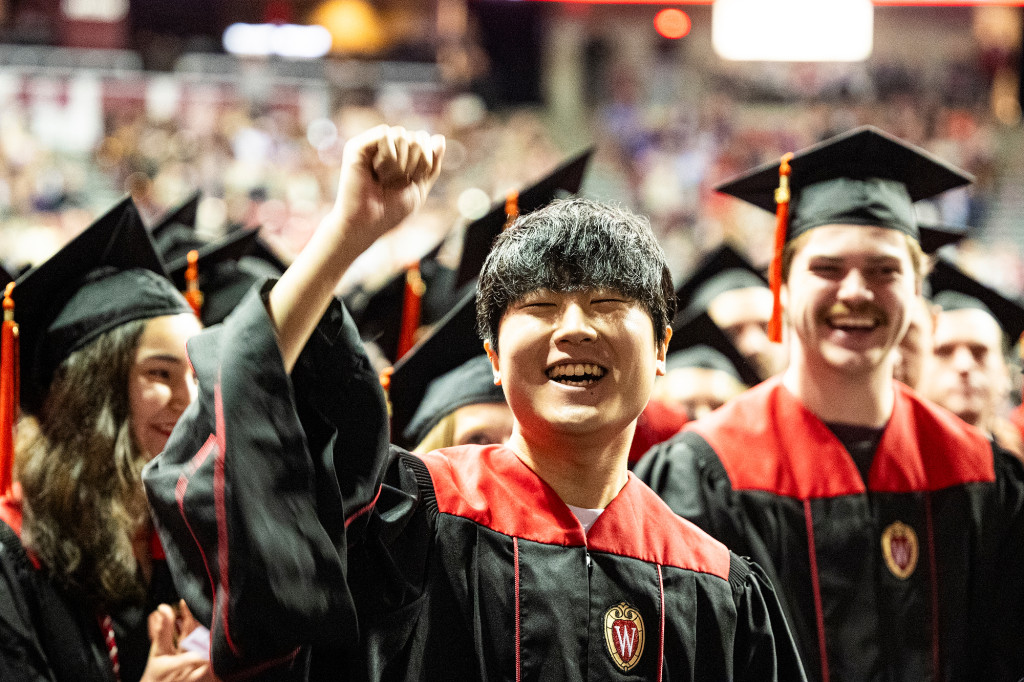 A man in a graduation gown stands and cheers.