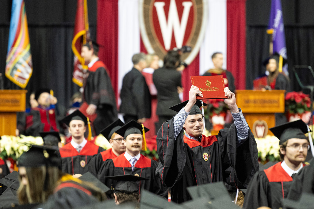 A group of people in black and red academic gowns smile and hold up diploma covers.