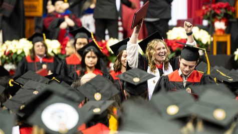 A group of people in black and red academic gowns smile and hold up diploma covers.