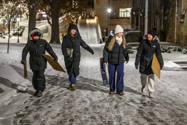 Four people carry cardboard sleds up a snowy hillside in the dark.