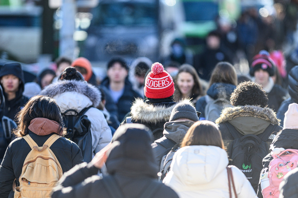 A group of pedestrians walks, including one with a red Wisconsin Badgers hat.