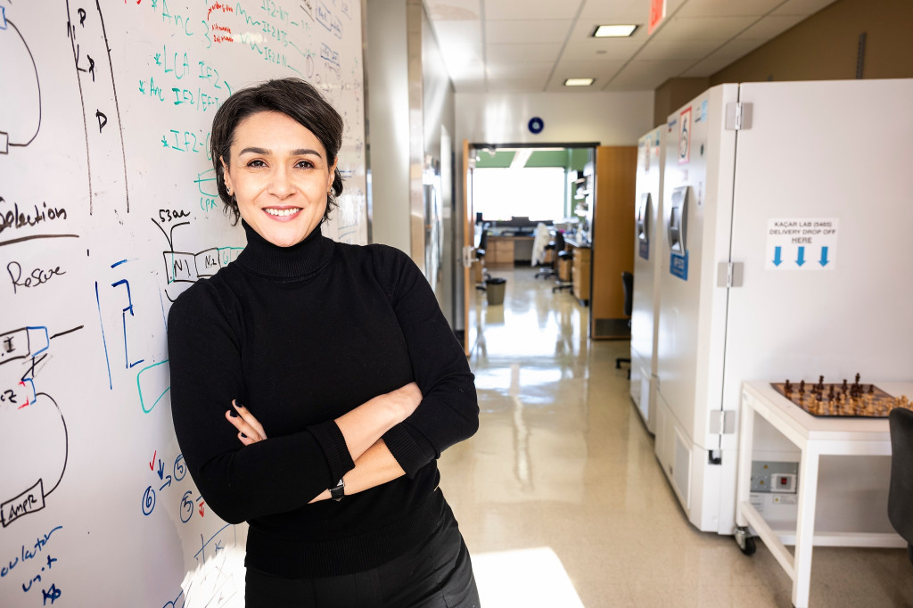 A woman leans against a white board filled with equations and sketches in a laboratory setting.