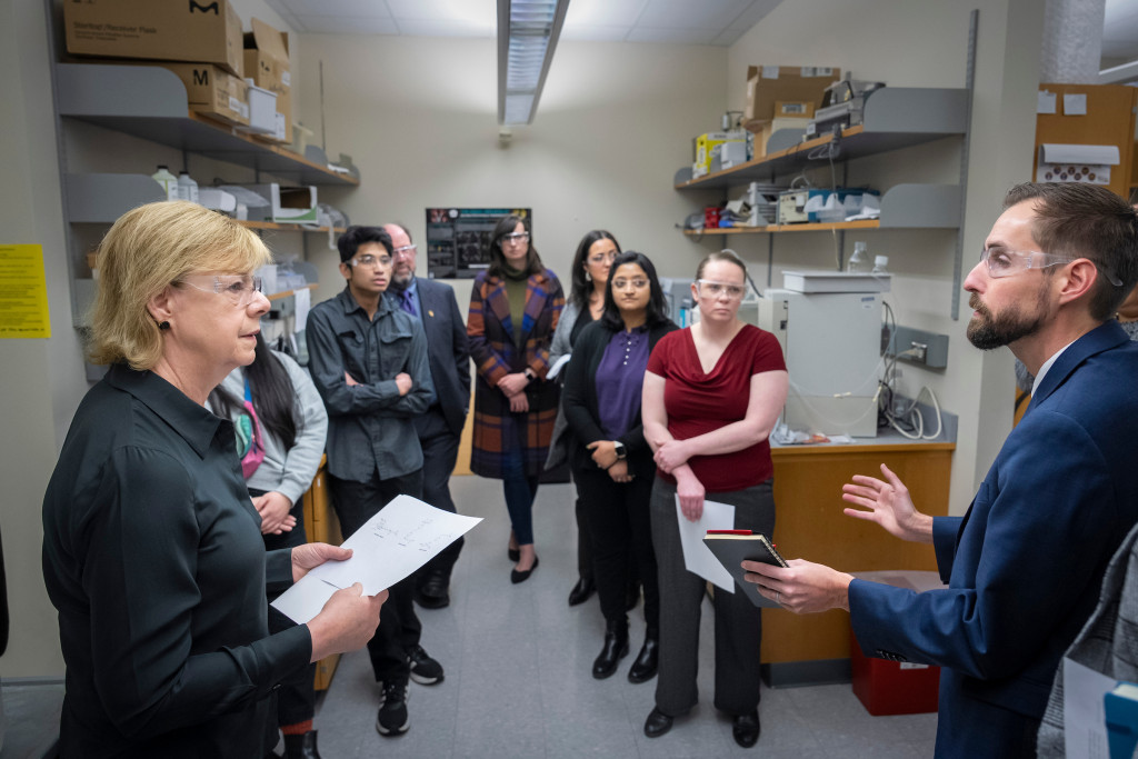 A small group gathers in a lab as part of a campus tour. One man addresses the crowd and answers questions about the space.