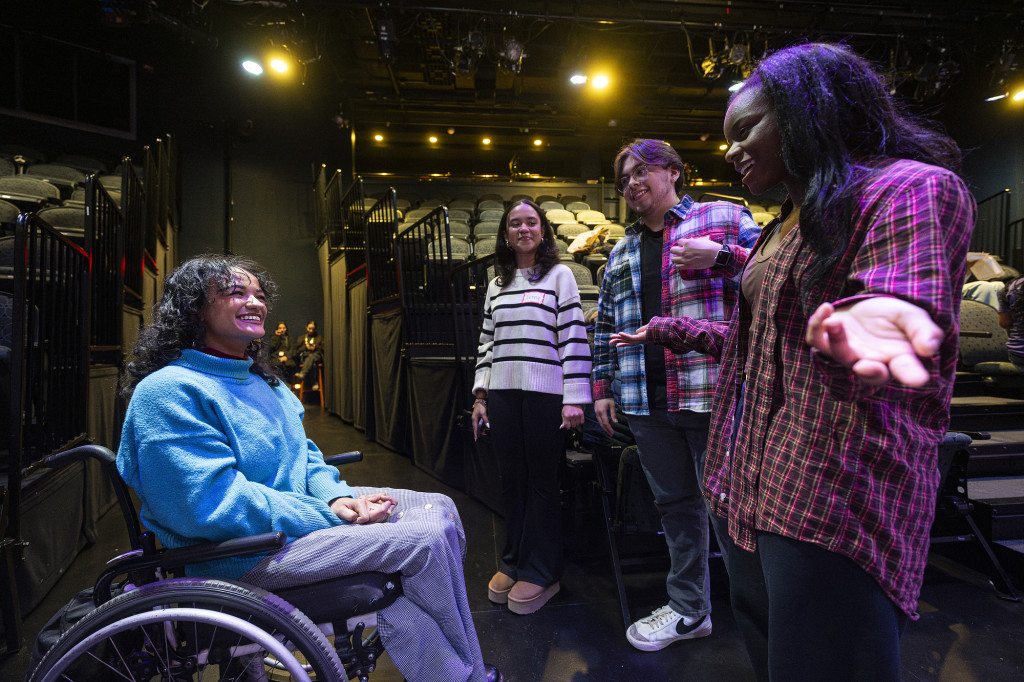 A woman in a wheelchair talks to several people on a darkened stage.