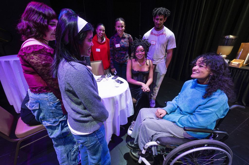 People gather on a darkened stage and talk to a woman sitting in a wheelchair.