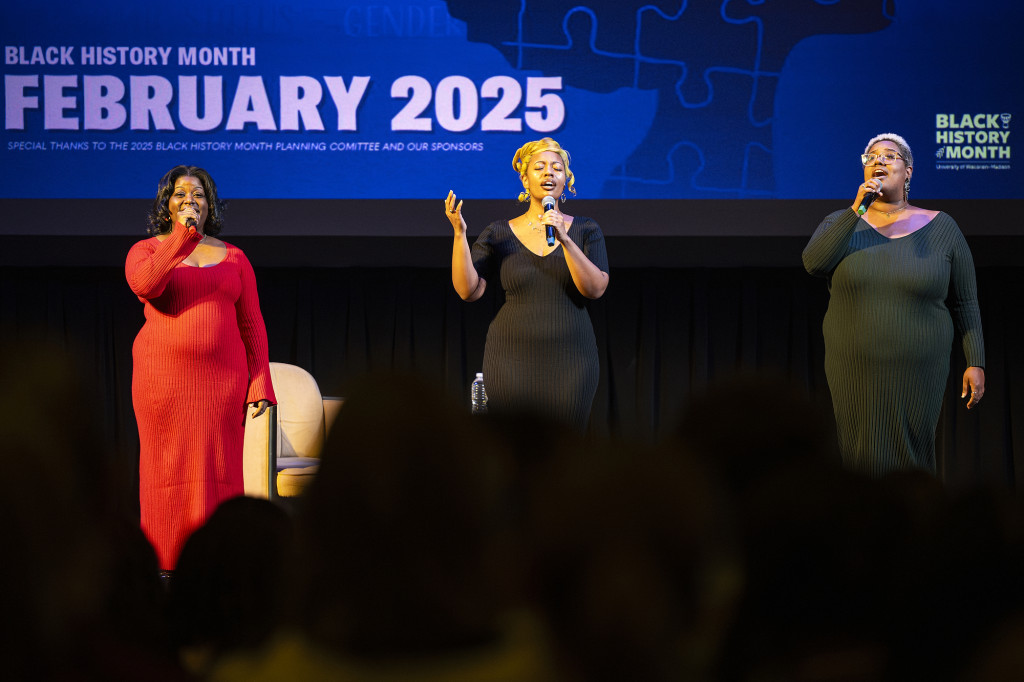 Three women stand on a darkened stage and sing.