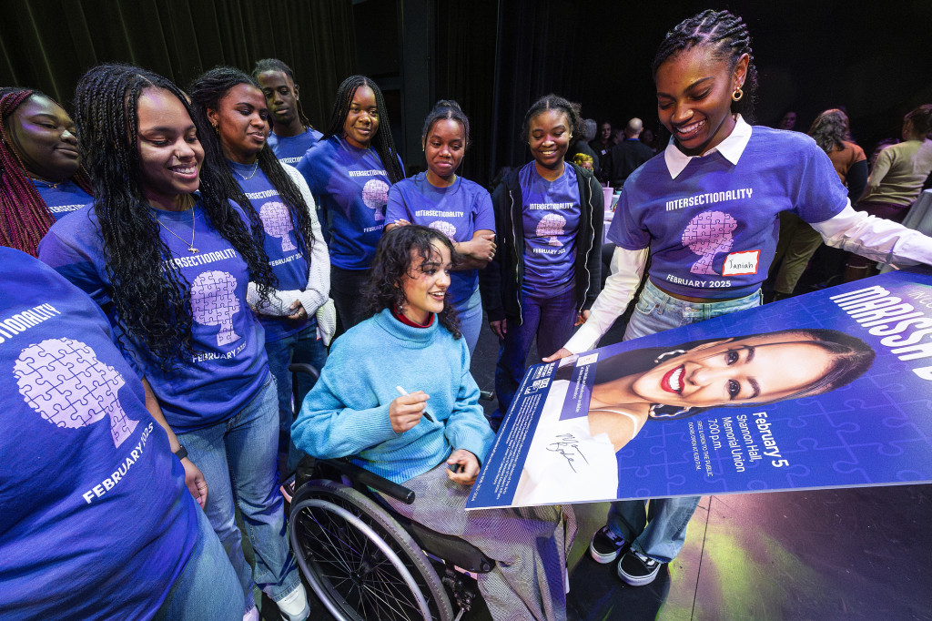 A woman in a wheelchair signs a poster while people are gathered around her.