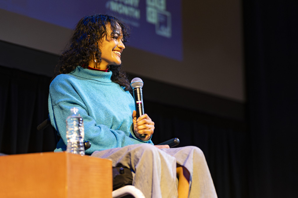 A woman in a wheelchair holds a microphone on a stage and smiles at the audience.