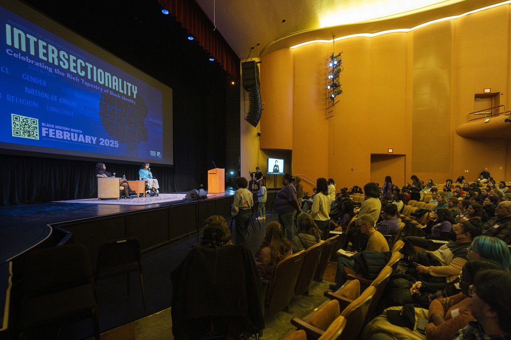 An overall view of a theater, with the stage on left with two people chatting on it, and the audience seated at right.