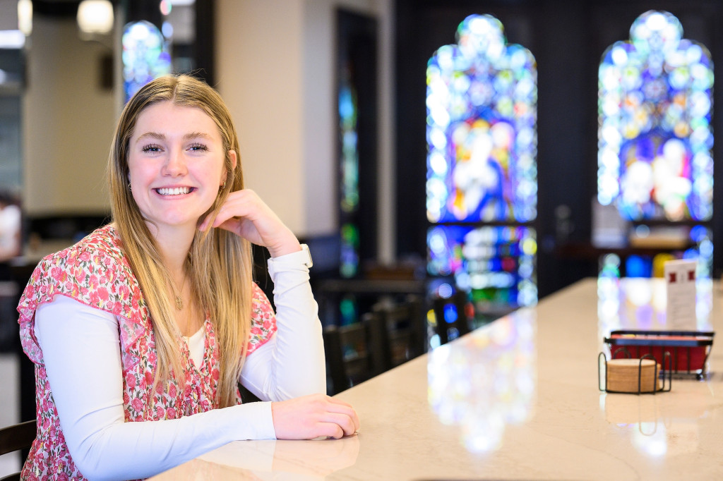 Brooke Brennan smiles to the camera for a portrait photo.She's sitting at a table in a large room with stained glass windows.