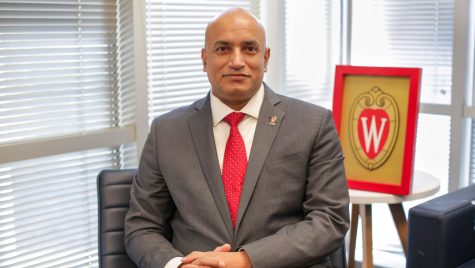 Portrait photo of Devesh Ranjan wearing a Wisconsin red tie and sitting next to a UW-Madison crest.