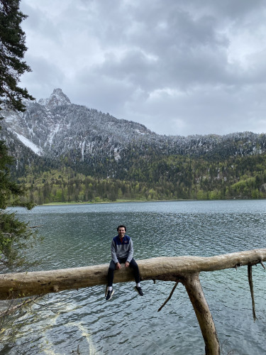 A person sits on a log that juts over a body of water, with a mountain in the background.