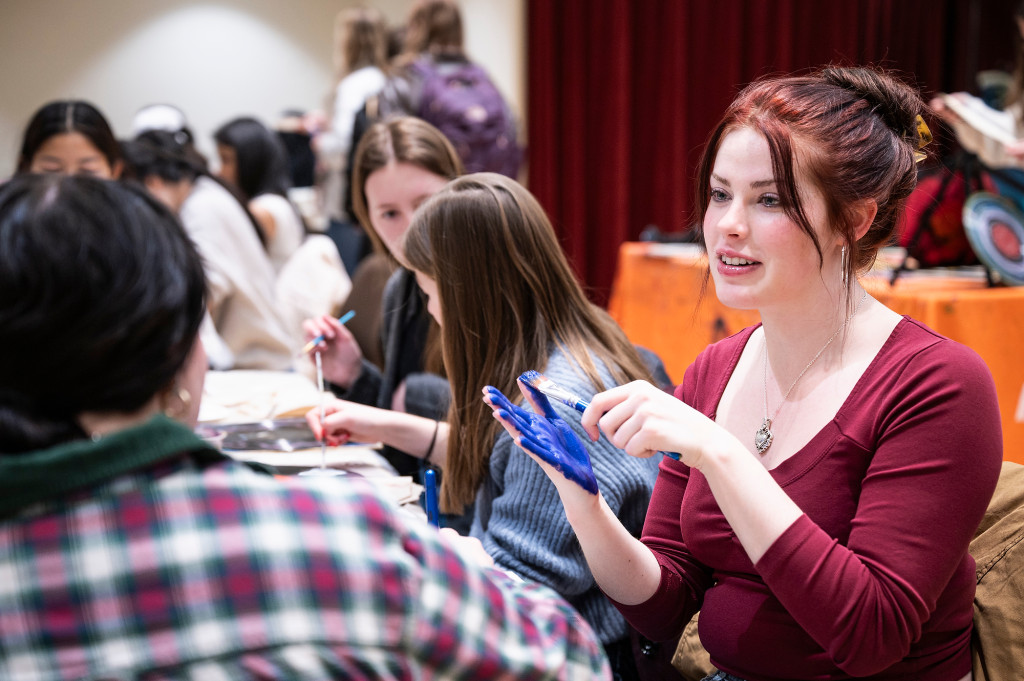 A woman paints on her hand with blue paint as she sits at a table where students are painting.