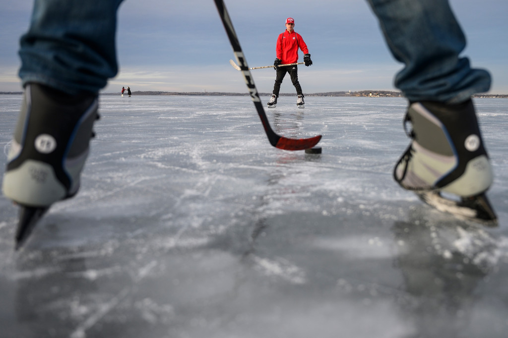 A photo shows a puck on a stick between someone's legs as another person with a stick prepares to catch a pass.