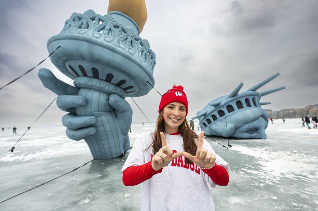 A young woman wearing a Wisconsin shirt holds her hands in a W formation in front of a rendition of the head and lamp of the statue of liberty, on an icy lake surface.