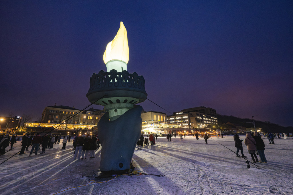 A torch recreating that held by the statue of liberty lights up a frozen lake, with people and lit buildings in the background.