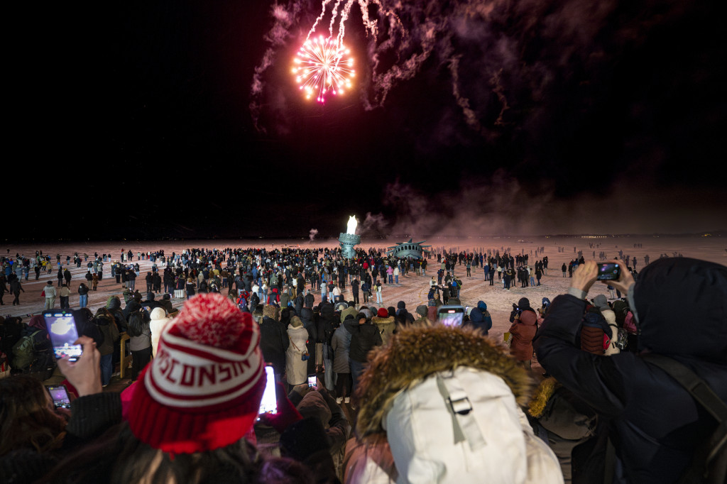 Fireworks light up the night sky above a frozen lake with people gathered on it, as well as a replica of the statue of liberty's head and torch.