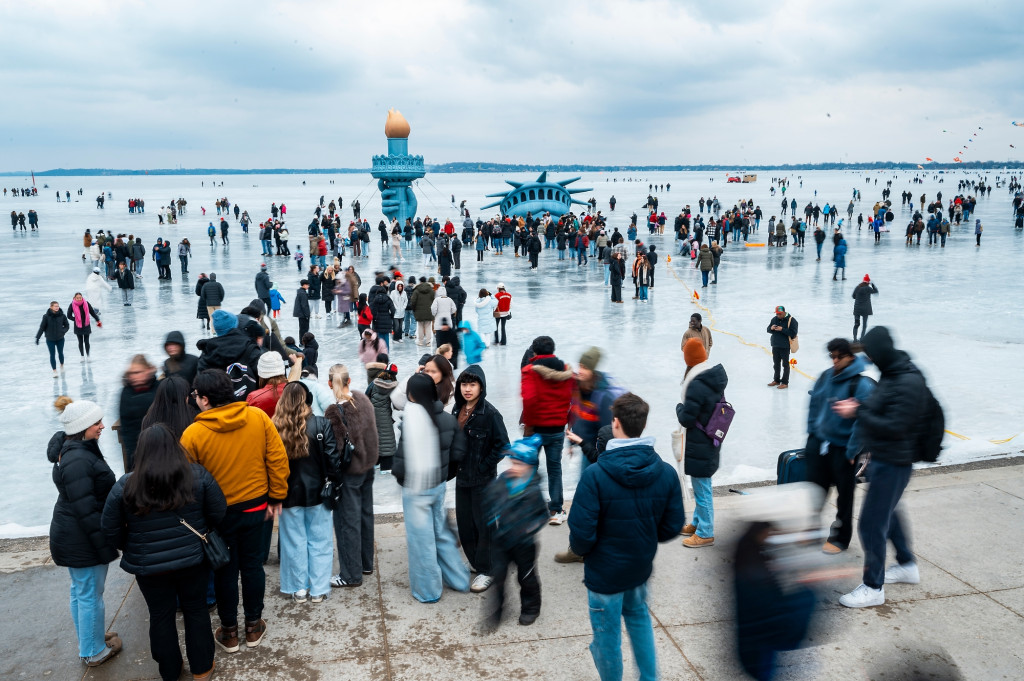 A crowd of people is shown on an ice lake around a replica of the State of LIberty's head and torch.