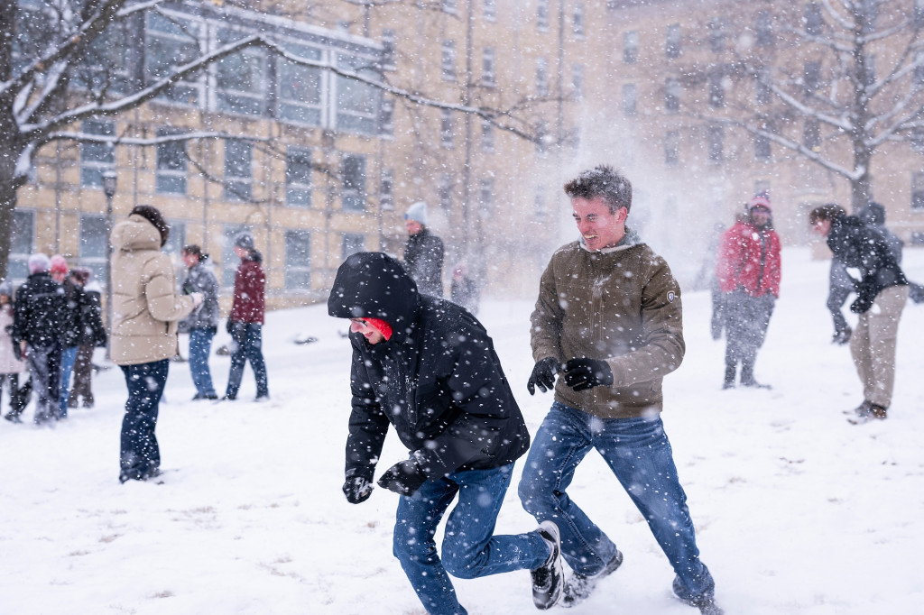 Two students are in the foreground of a group of students. All are laughing and throwing snowballs at each other on Bascom Hill.