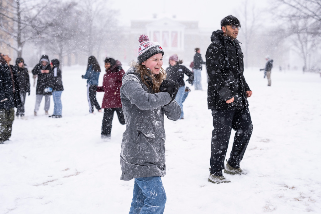 A student at the center of the picture gathers snow in her hands as she shapes a snowball. Laughing, she looks at her friends who are off camera.