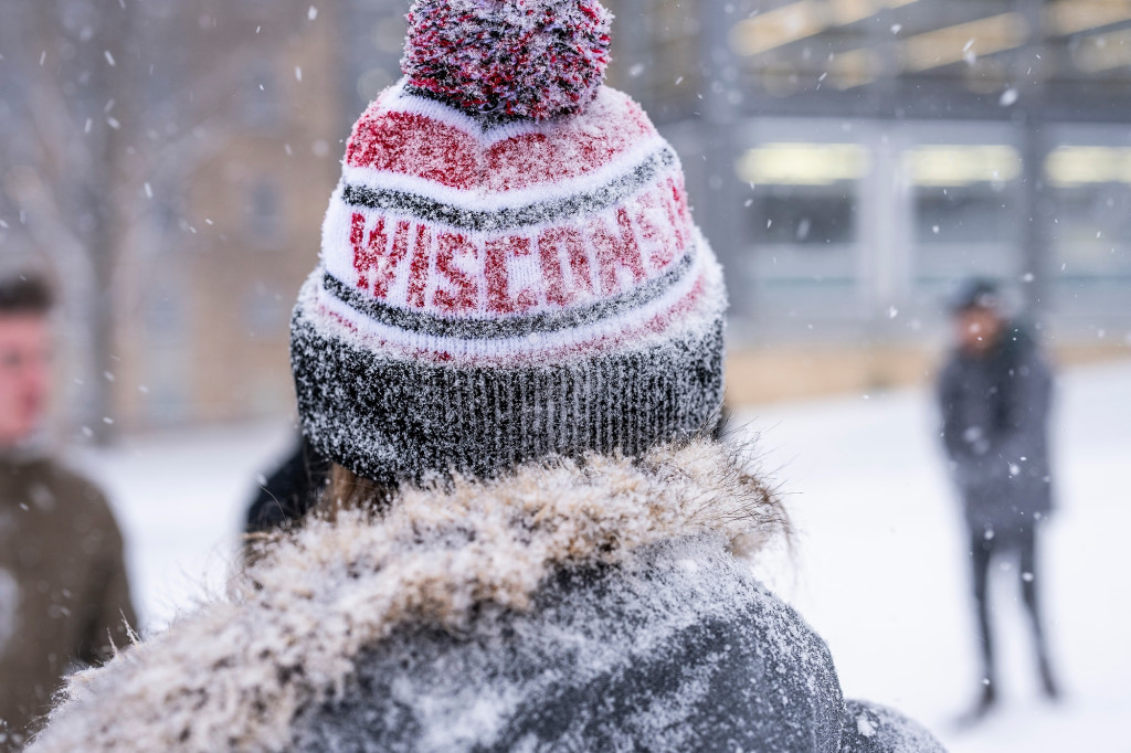 This closeup shows a person wearing a Wisconsin-branded winter hat that is covered in snow from a recent snowball fight.