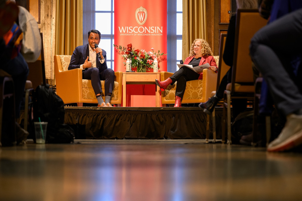 Two people sitting on stage in armchairs talk.