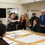 A professor stands at the end of a table filled with screen prints. She points to a print at the other end of the table as she discusses her classes with Mnookin and Governor Evers, who stand at the side of the table.