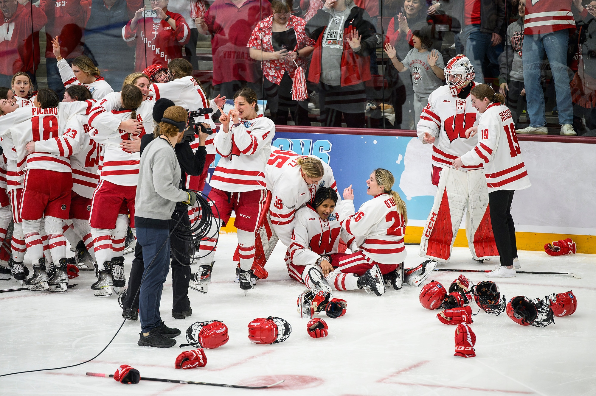 The Wisconsin Women's Hockey Team celebrates on the ice after winning. Some players embrace while others sit on the ice in disbelief.