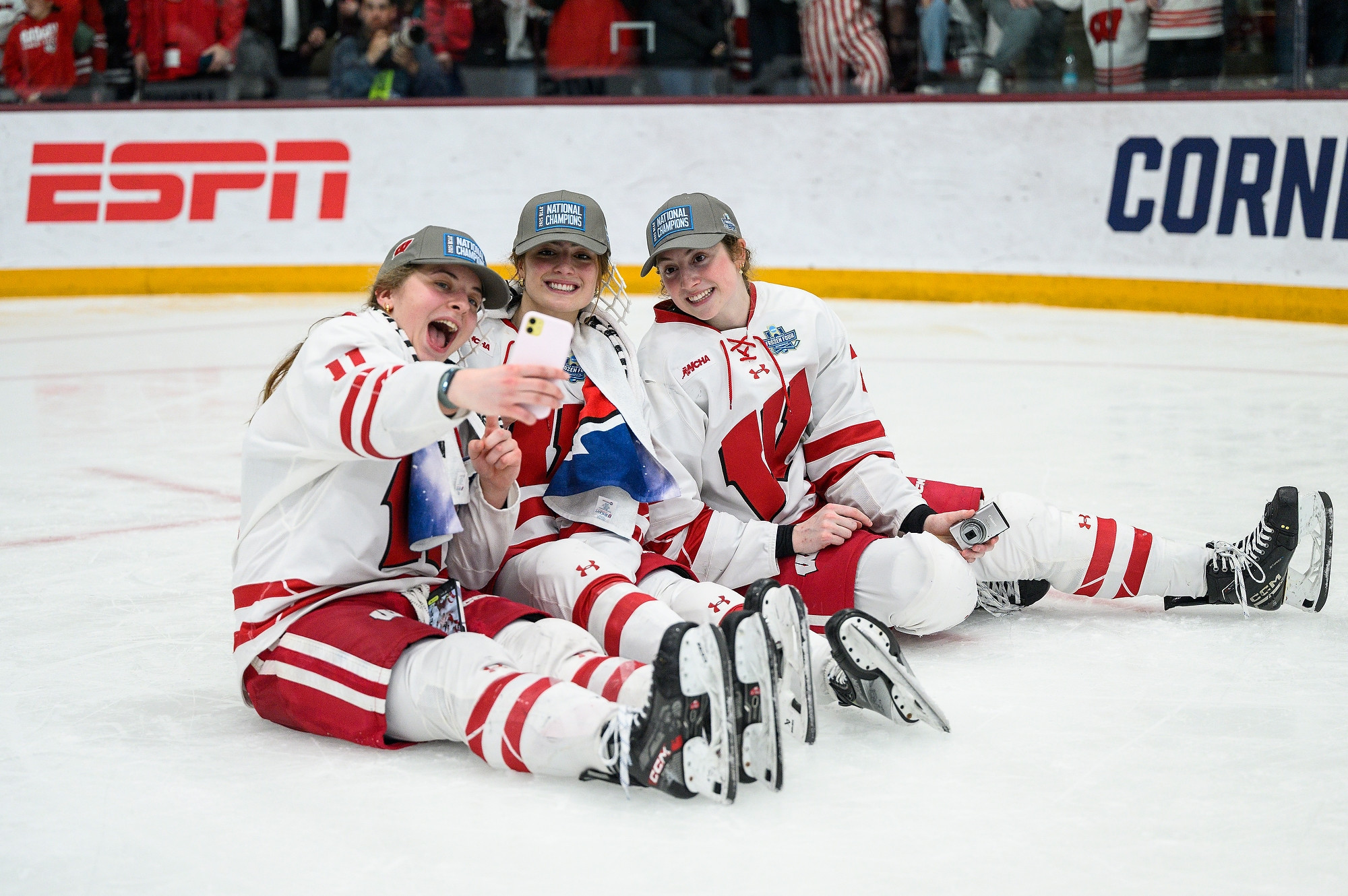 Three Badger players wear their uniforms and newly adorned National Champions hat while sitting on the ice together. They look at the phone one of the players holds up for a photo.