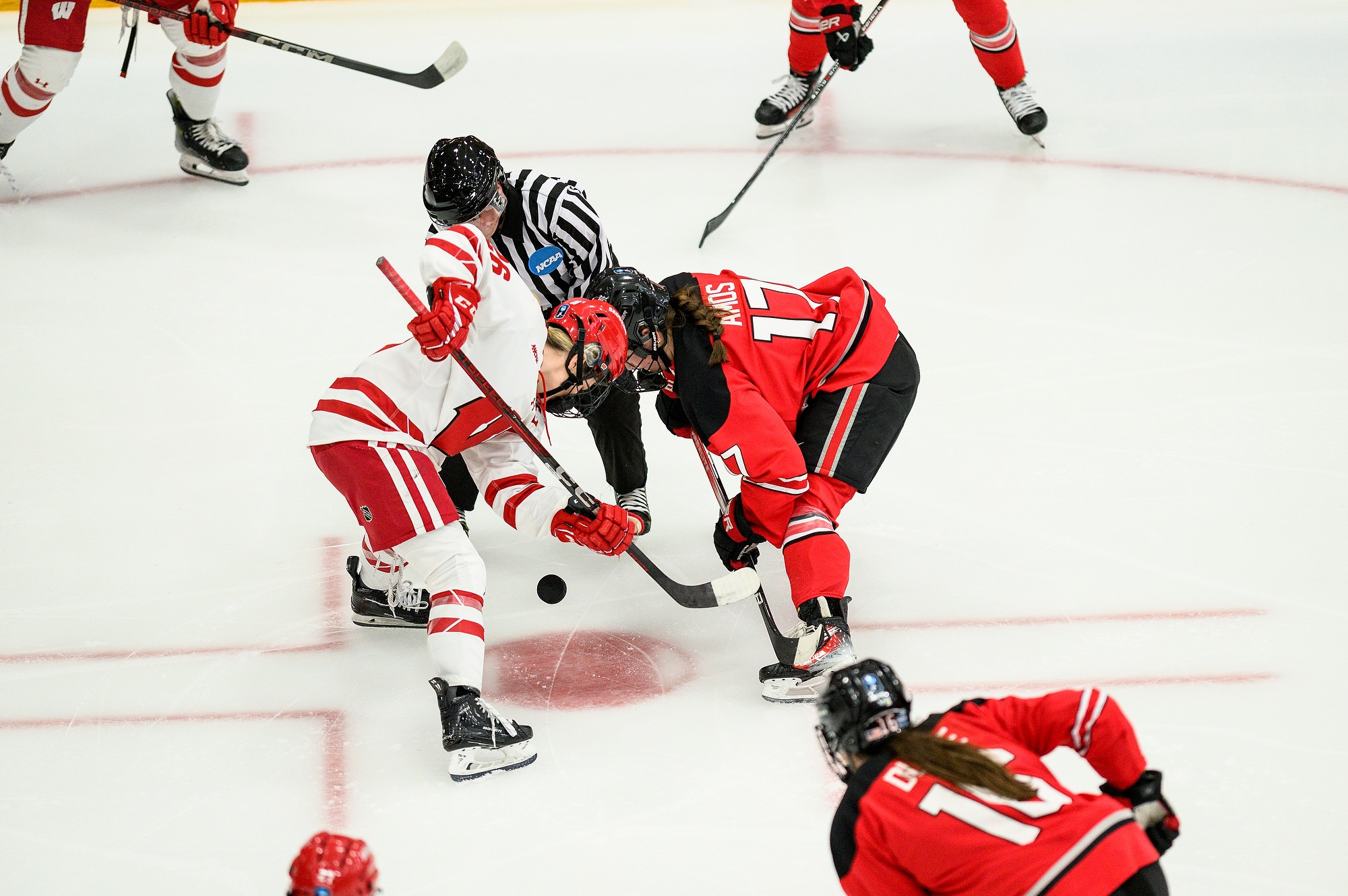 The camera focuses on two women's hockey players on opposing teams, standing at the center of the ice ready for the puck to drop from the referee's hands.