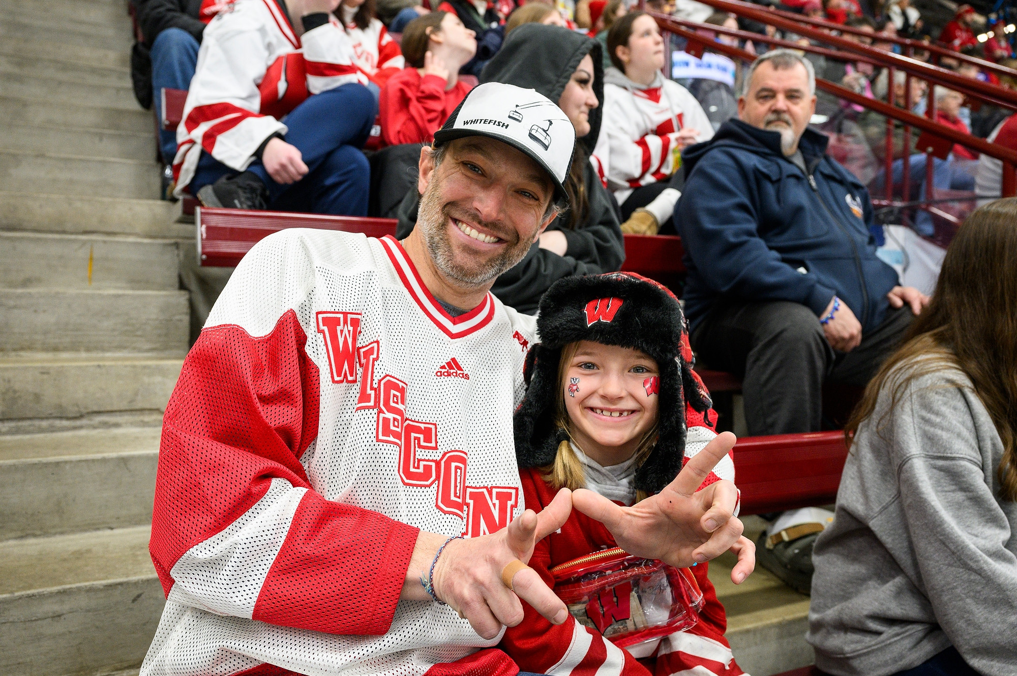 A man wearing a Wisconsin Badgers hockey jersey sits close to his daughter. His arm is wrapped around her as he flashes a "W" sign that he makes with his two hands. The daughter wears a Wisconsin Badgers bomber hat and flashes a smile at the camera.