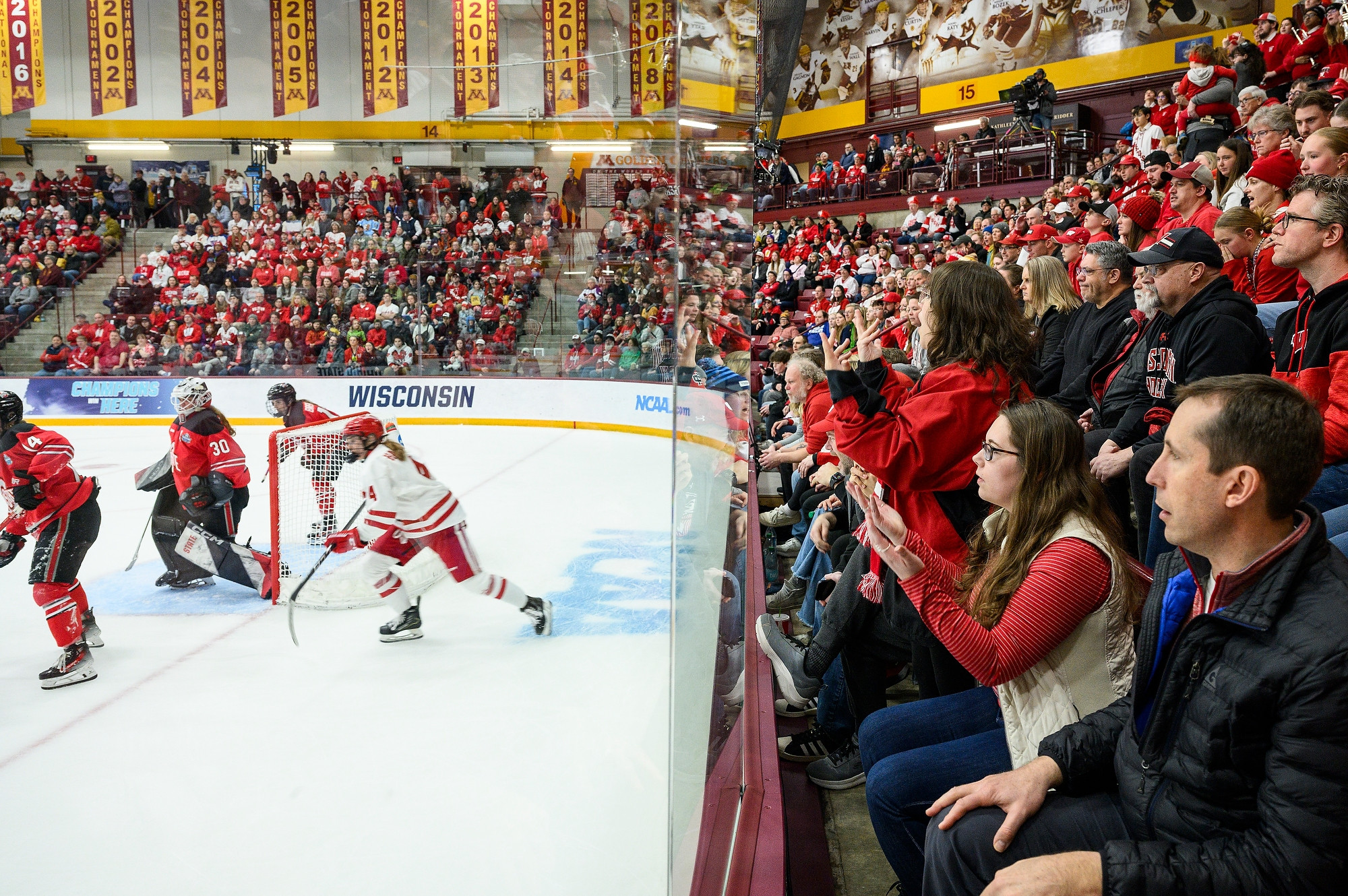 A crowd of mostly Badger fans encircles the hockey rink. The fans react to the matchup unfolding on the ice on the other side of the protective glass.
