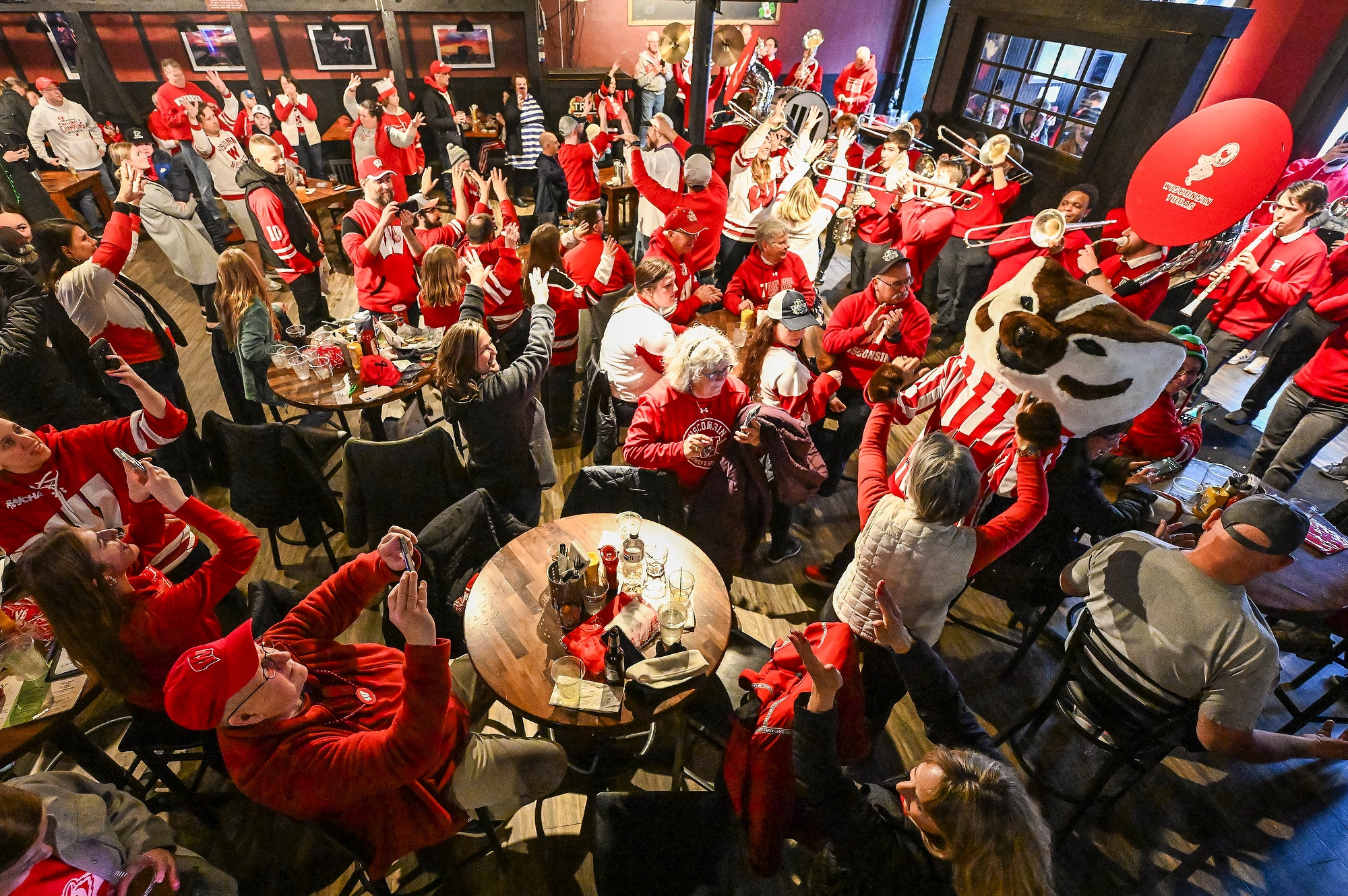 An overhead photo of a large group of Badger fans filling a local restaurant. The fans wear red and white attire and sing along to the UW Marching Band and Bucky Badger.