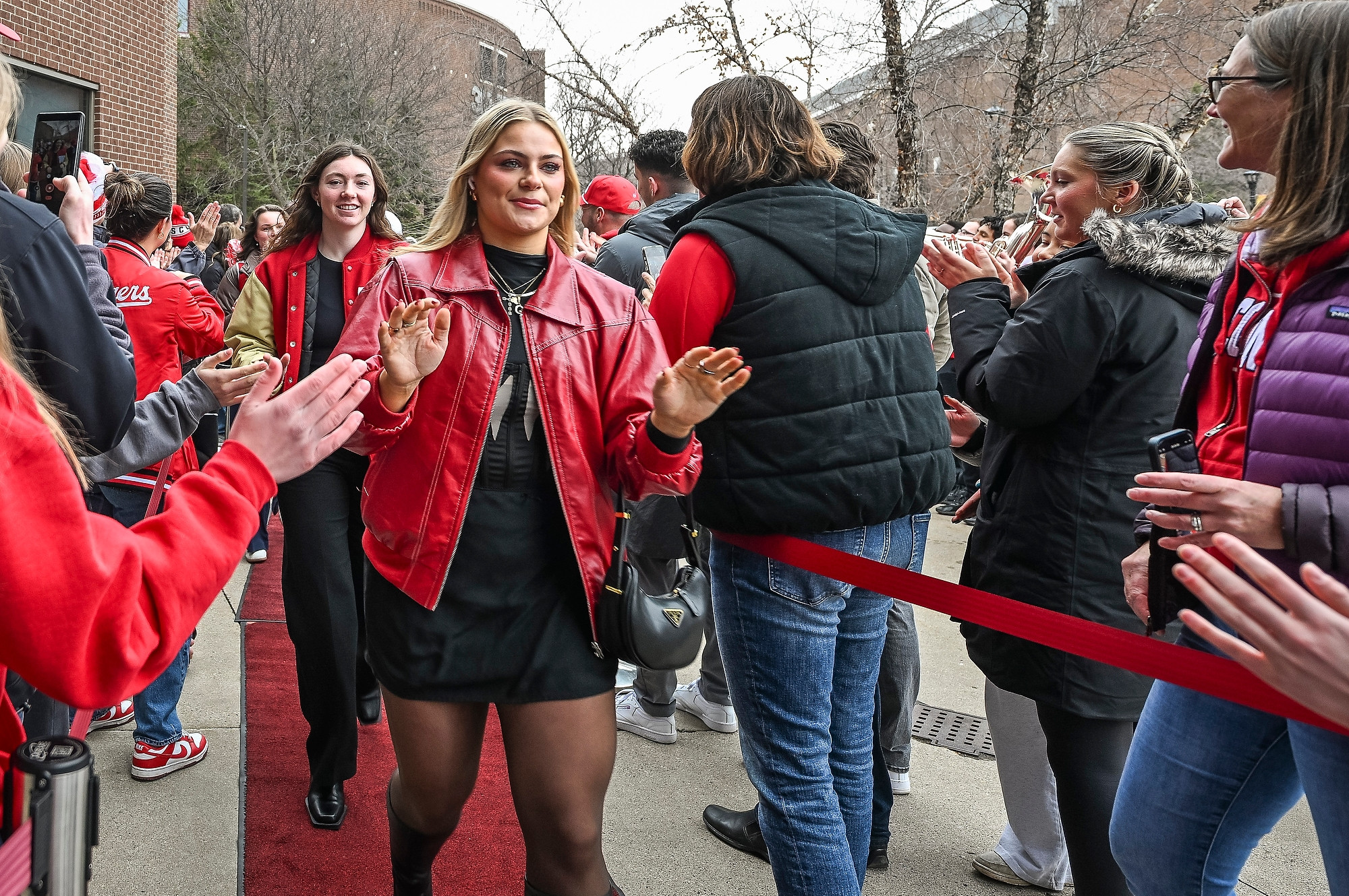 A crowd of fans stand on either side of a red carpet that leads to the entrance of the hockey arena. Members of the Badger women's hockey team done semi-formal attire as they follow the carpet inside, giving high fives to fans that they pass by.