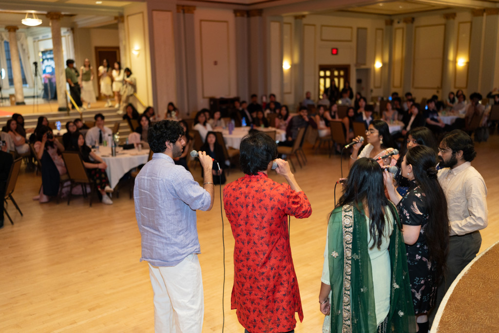 Seven student wear cultural attire and stand in a semicircle in front of a large audience. They each sing into a microphone they are holding as they perform for the room.