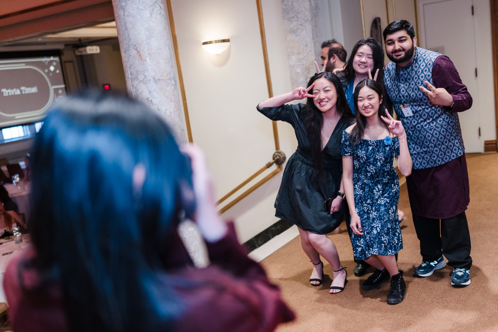 Four students pose with their fingers formed into peace signs and smile as a fellow student stands in the foreground of the picture holding up a camera and taking a photo of the group.