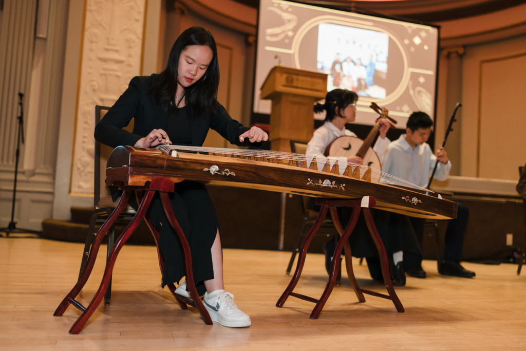 A students sits and play a traditional Asian musical instrument next to fellow bandmates who join her on stage and are pictured in the background of the photo.