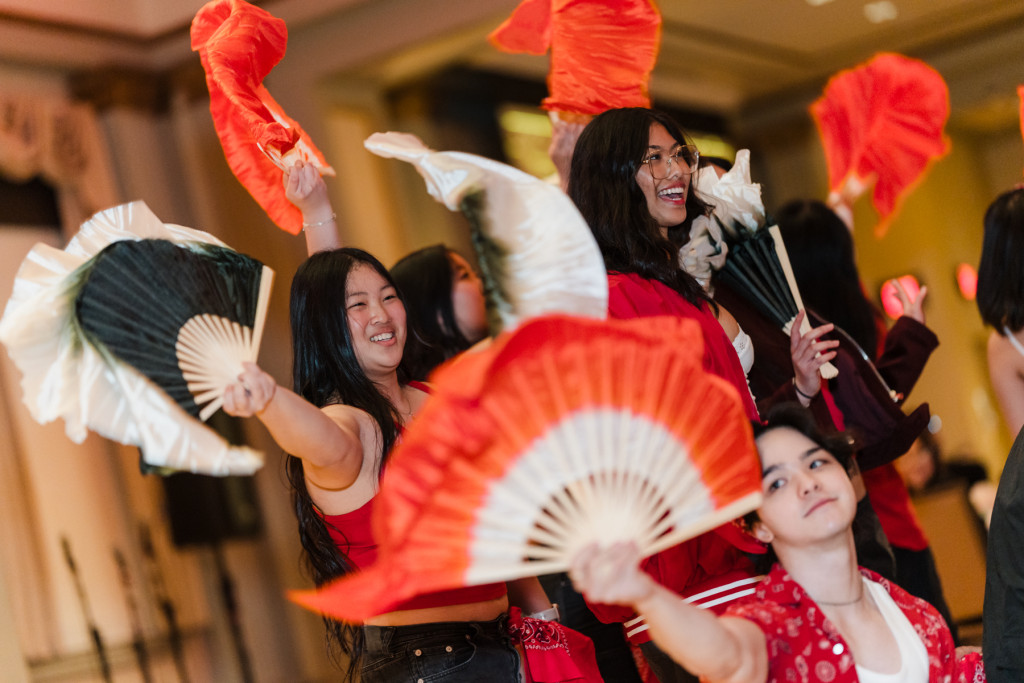 A group of students cluster together for a pose at the end of a dance performance. Each wave black and white or red and white flags as they smile to the audience that is not pictured.