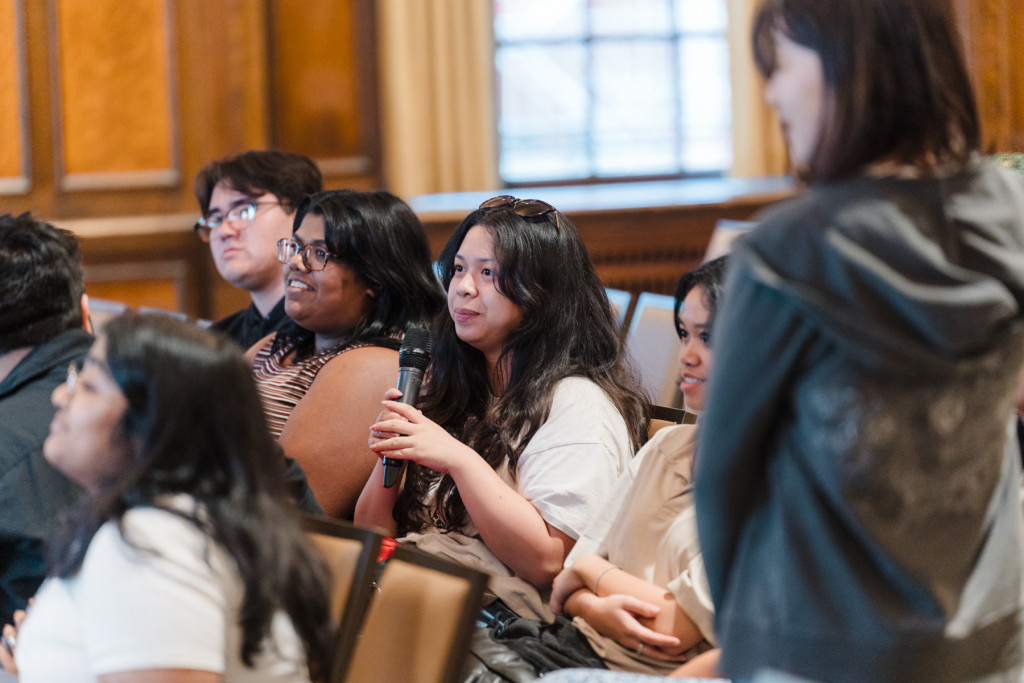 Students sit in chairs facing the table at the front of the room. The photo shows a woman holding the microphone in her left hand as she asks Perez a question.