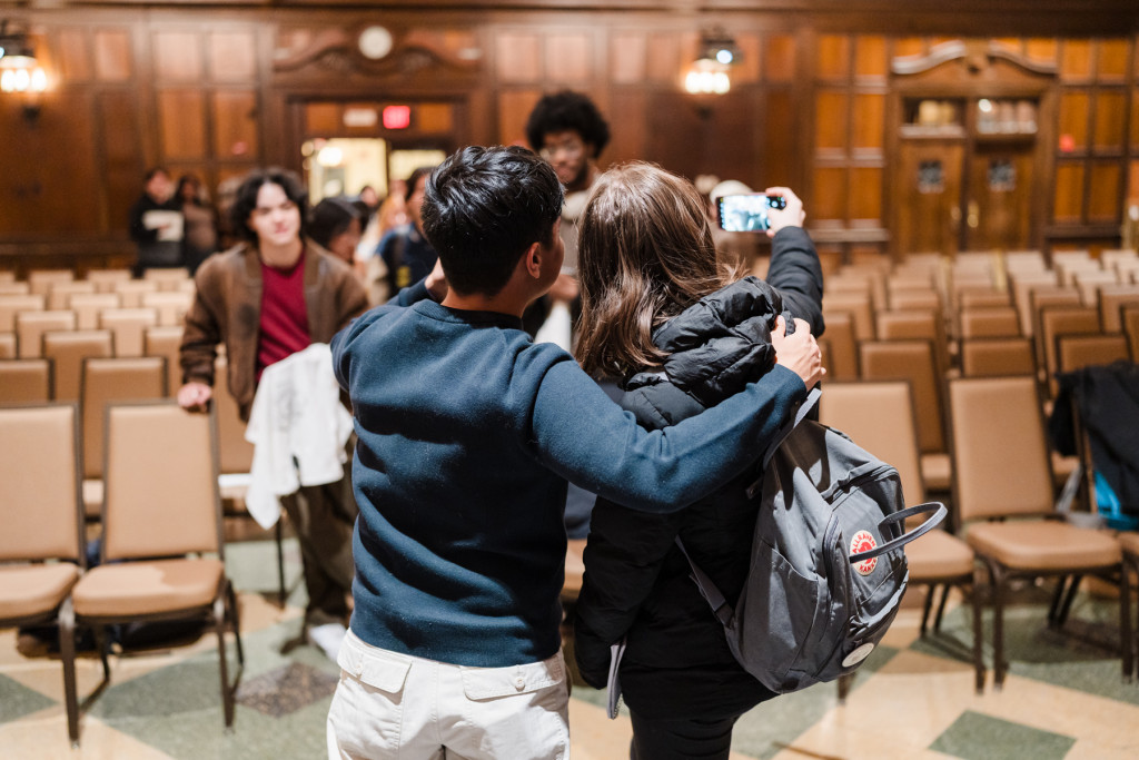 Seen from behind, Perez hold his arm around a student as they hold up a camera for a selfie. Students wait in the audience for their turn to speak to Perez.