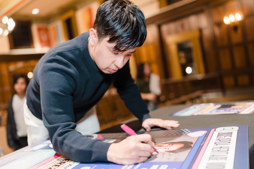 Perez stands over a table with a marker in his hand. On the table is a large event poster with his photo, which he signs.