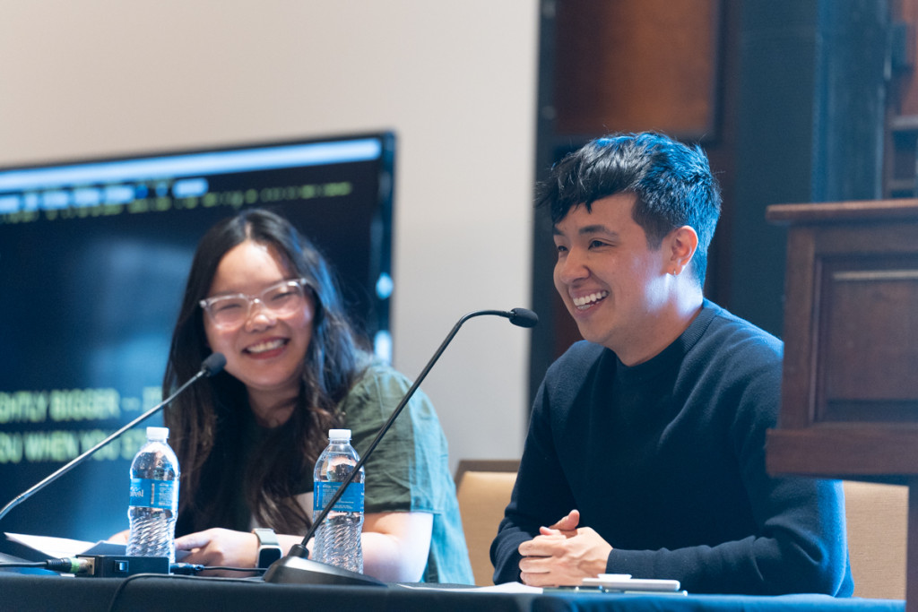 Dong and Perez sit at a table. Dong smiles while Perez speaks into a table top microphone positioned in front of him.