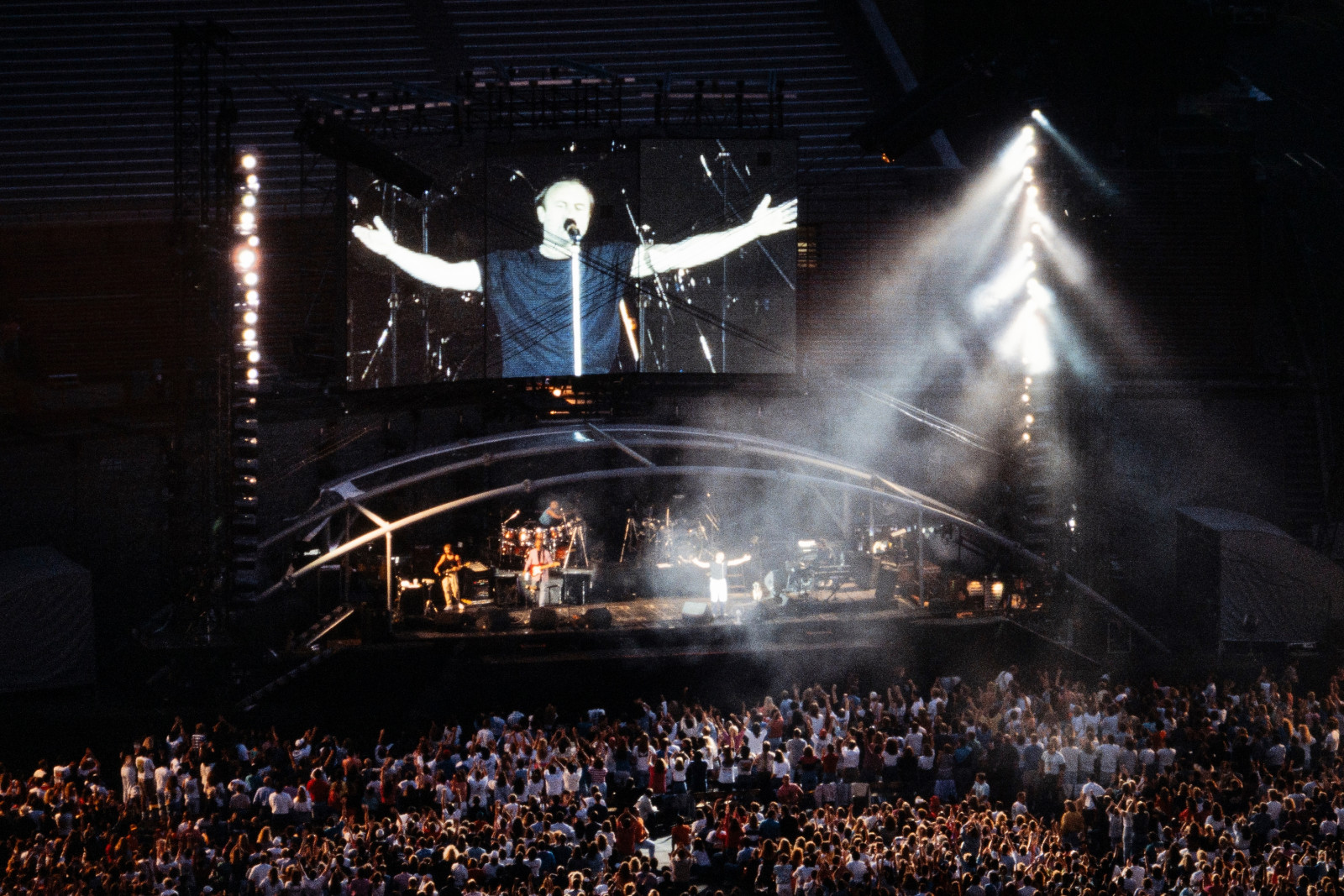 An overview angle of a Genesis concert with tightly packed fans and the band members on stage, with a large video screen showing lead singer Phil Collins singing and holding his arms open.