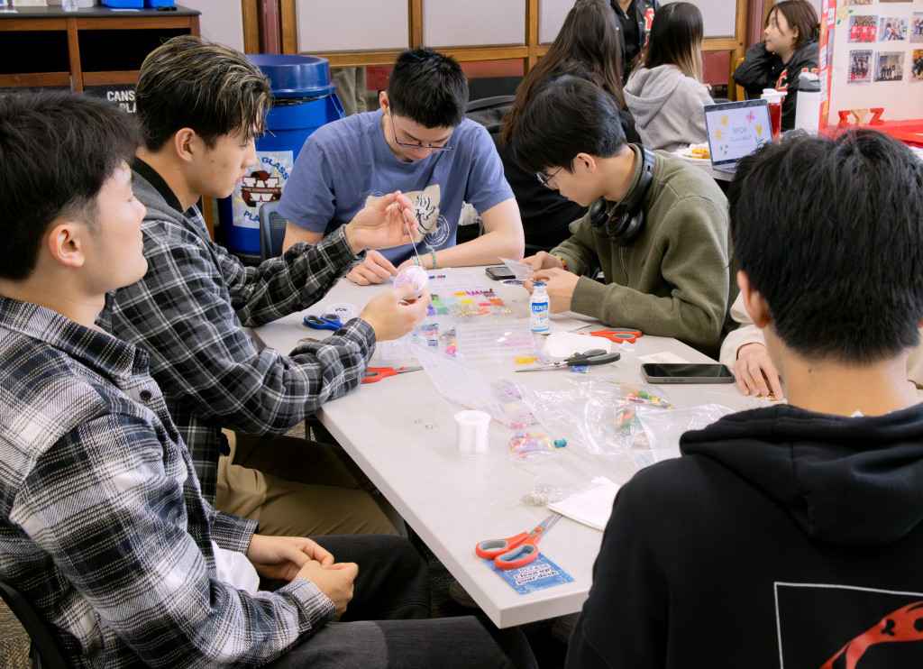 Five students sit around a rectangular table. They focus on the craft they are each working on surrounded by bags and containers fulled with different color beads.