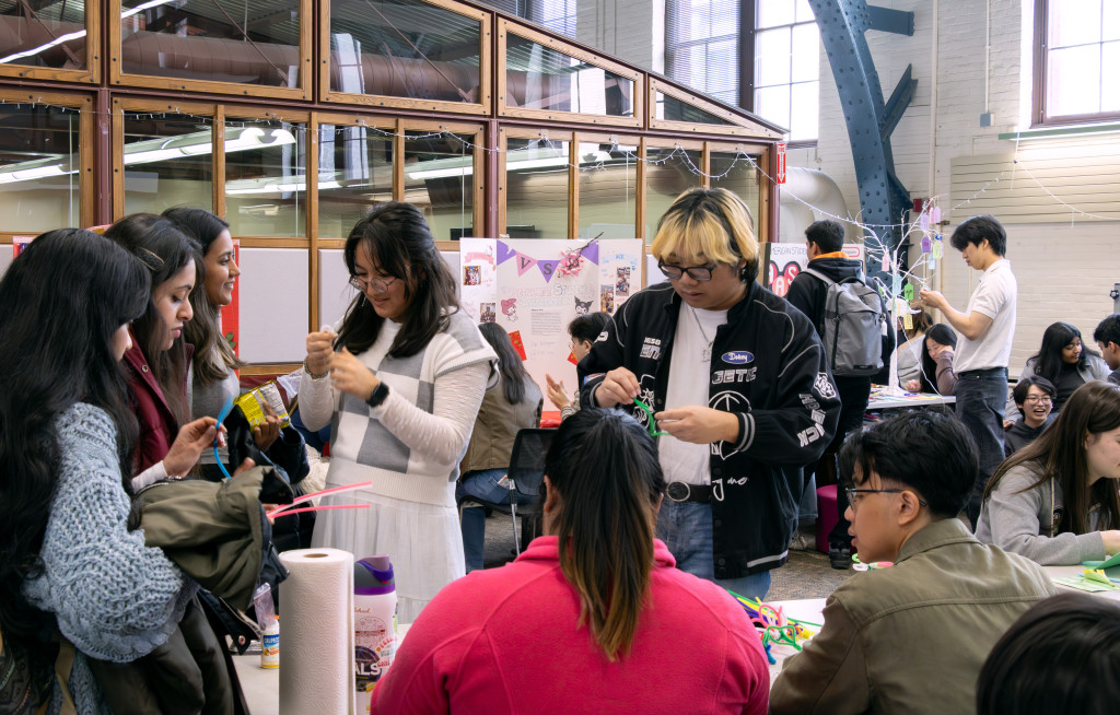 A group of students stand around a stable, which is staffed by two fellow students. The students who stand mold pipe cleaners into different floral shapes.