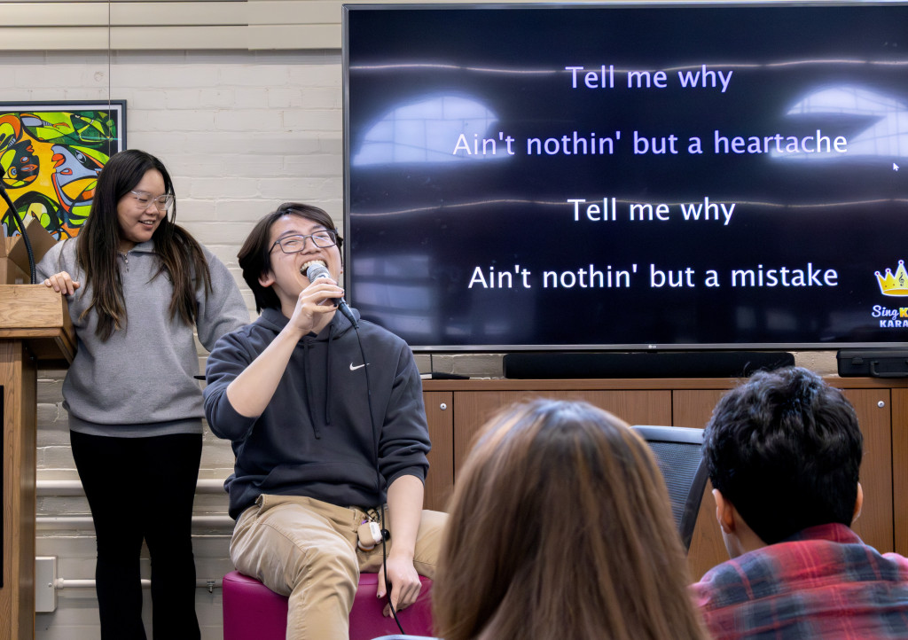 Xie sits on a stool in front of a large TV screen showing karaoke lyrics to the Backstreet Boy's song, 'I want it that way.' Xie can be seen singing into a microphone.