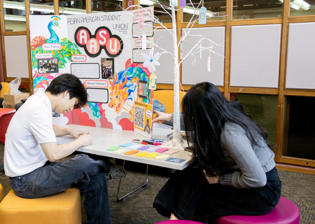 A small white tree sits on a table with small hand-written messages hanging from the branches. Two students sit at the table writing their own messages on pieces of paper that they will then hang on the tree with the other wishes.