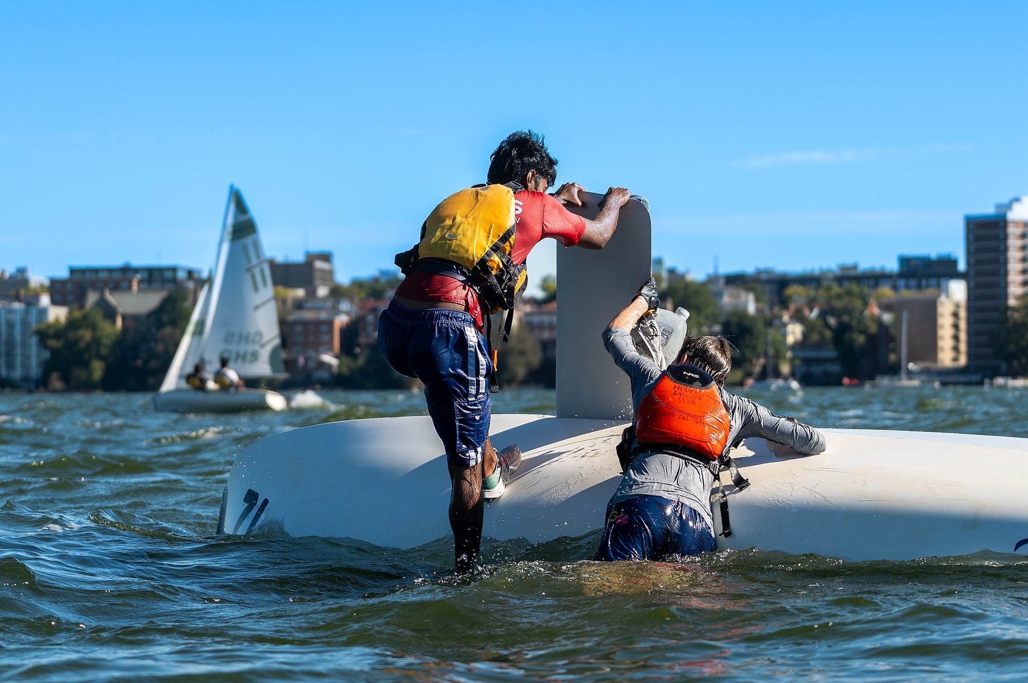 Two people cling to the hull of an overturned sailboat.