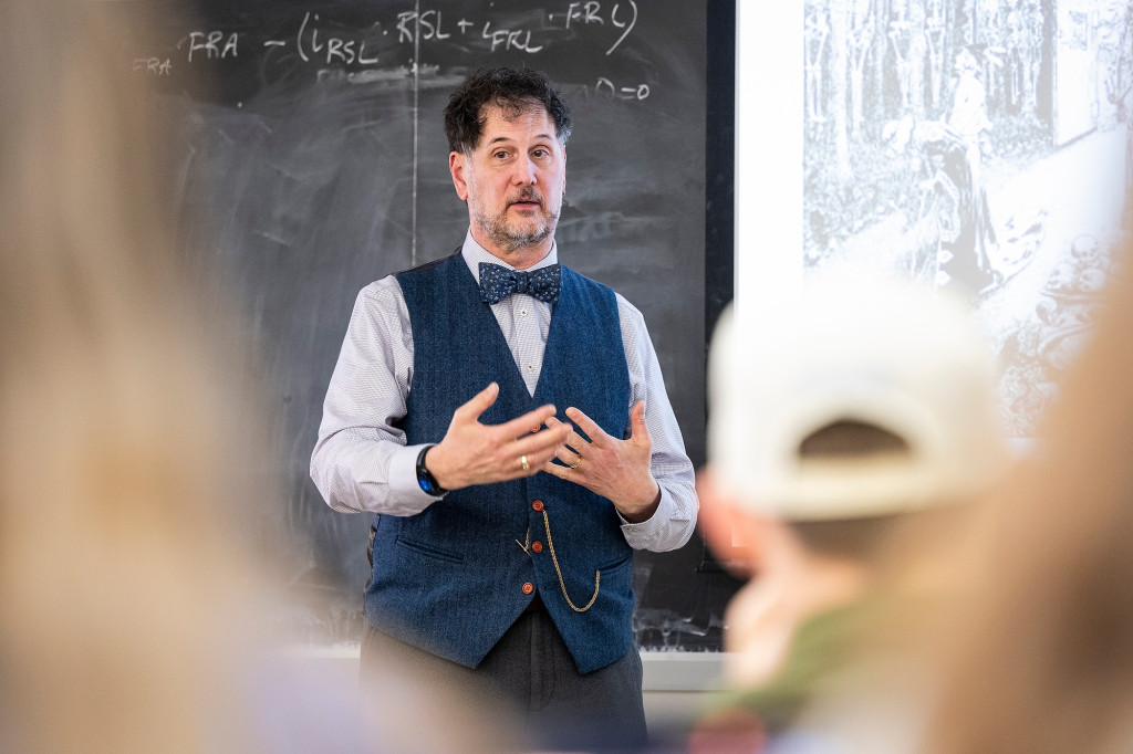 A man stands in front of a chalkboard and speaks to a class of students.